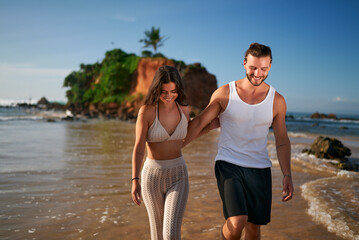 Happy couple walks barefoot on tropical beach at sunset, man leads woman by hand near ocean, romantic stroll by sea, young love on summer holiday, relaxed lifestyle, travel, vacation mood.