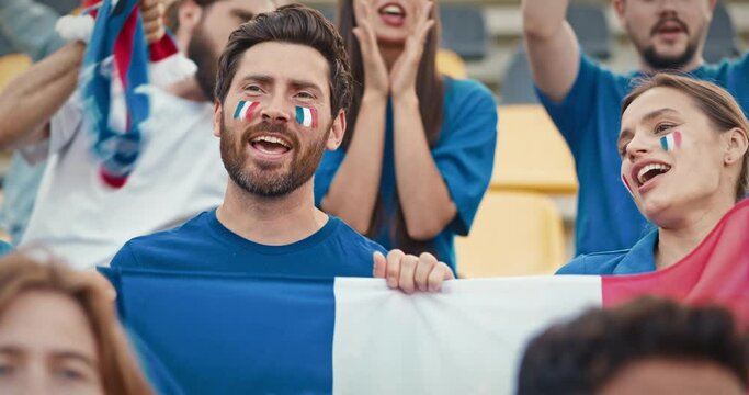 Cute Caucasian couple singing and shouting something from stadium tribunes. Fans holding French national flag and jumping with it. Painted French flags on their faces to show support.