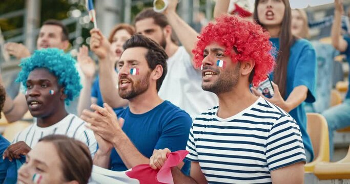 Group of multi-ethnic friends wearing wigs and French flags painted on their faces. Cheering for France from stadium tribunes. Shouting words of support. Singing some songs. Having good time.