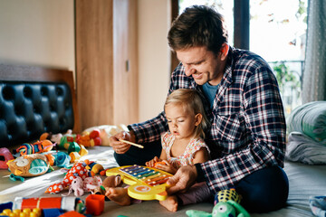 Dad plays with a little girl on a colorful xylophone, sitting behind her on the bed