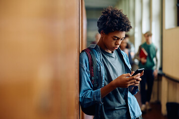 Black student texting on mobile phone in hallway at high school.
