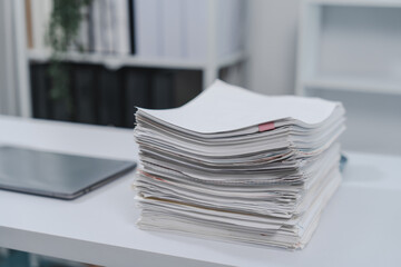 stack of assorted papers and documents with a red pen on top, situated on a clean, white desk, with a bookshelf and green plant in the background.