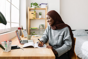 Young adult african student woman taking notes while using laptop computer at home. Millennial black female in muslim headscarf learning online listening virtual video call. Education concept.