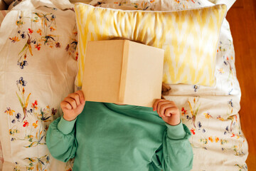 Caucasian blond teenage girl is lying on the bed and reading book.