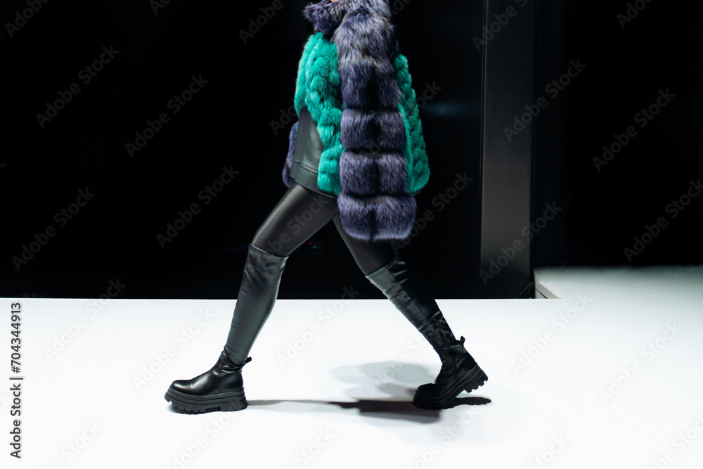 Wall mural female figure dressed in a fur coat and black leather boots walking on black background.