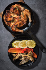 Fried chicken on a black frying pan with vegetables on a grey background.