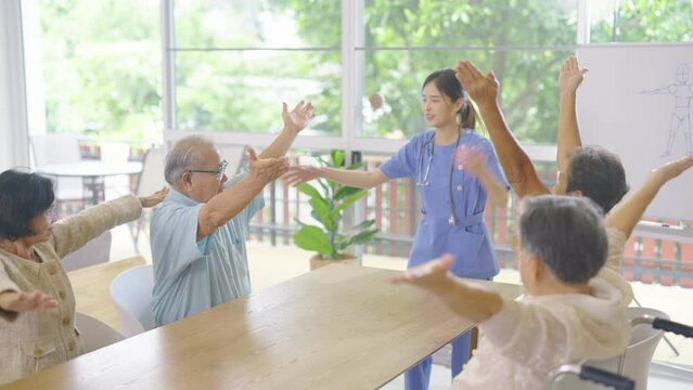 Side view young Asian nurse stand in front of group of senior or elderly people to show as example for exercise hands in room of clinic or healthcare center.