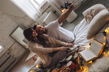 Happy young couple relaxing together in the bedroom at home, taking pictures. Man and woman enjoying lazy cozy weekend at home, embracing, kissing, cuddling. Simple pleasures, domestic life - obrazy, fototapety, plakaty