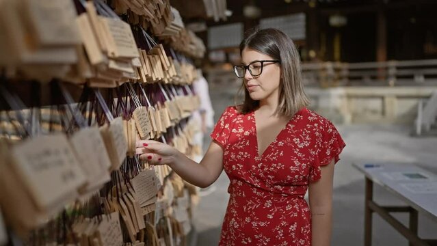 Portrait of a beautiful hispanic woman in glasses at japan's iconic meiji temple, affectionately admiring the japanese wooden ema prayer boards and soaking in the rich traditional culture