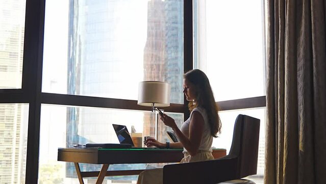 Stylish businesswoman freelancer working with laptop and smart phone, she is sitting on chair, enjoying panoramic view in city background. Low key photo