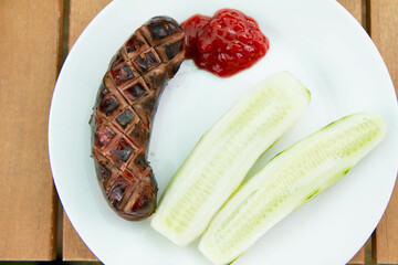 Barbequed Sausage served on a plate in a garden with ketchup and fresh cucumber - 704377310