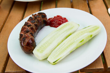 Barbequed Sausage served on a plate in a garden with ketchup and fresh cucumber - 704377313
