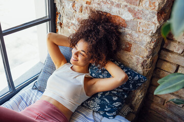 Pleased relaxed African American ethnicity girl in homewear sitting on windowsill