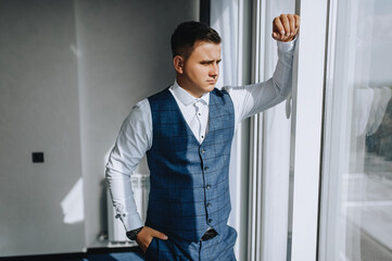 Stylish groom in a blue suit, vest stands near the window indoors, hotel in the morning. Wedding portrait, photography.