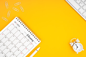 February 2024, Monthly desk calendar for 2024 year and alarm clock with paper clips on yellow color background.