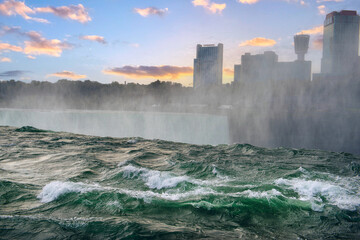 An unusual view of Niagara Falls from the very edge of the cliff. View of skyscrapers on the other side of the waterfall in Canada and the sunset sky.