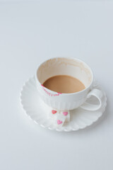 There is coffee with milk in a white coffee cup and saucer. There are lipstick marks on the cup. refined sugar with hearts in a saucer. White background