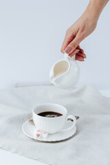 A woman's hand pours cream into coffee from a white milk jug into a white cup, next to it is refined sugar with hearts on a linen napkin. white background