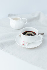 black coffee in a white cup with a saucer, next to it is refined sugar with hearts on a linen napkin. behind is a white milk jug on a white background