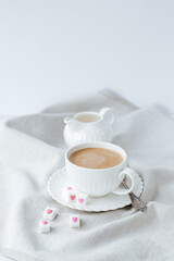coffee with milk in a white cup with a saucer, next to it is refined sugar with hearts on a linen napkin. behind is a white milk jug on a white background