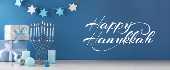Happy Hanukkah. Menorah, candles, dreidels and gifts on white table against blue background, banner...