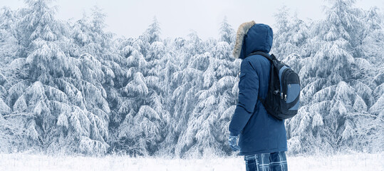 Back view of traveler in a blue jacket with a fur hood and a backpack on the background of a winter landscape during snowfall