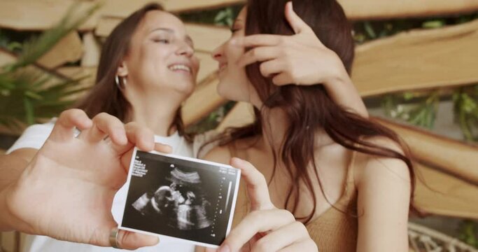 Happy lesbian women in love, expect baby and showing ultrasound picture while sitting embracing. Joyful pregnant female in same sex marriage. Maternity concept. LGBT relationship