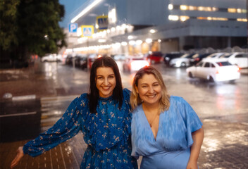 Fototapeta premium Two young women in elegant dresses hug and laugh in rain in evening
