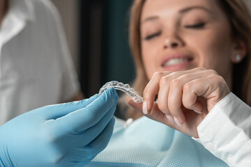 Attentive dentist demonstrating aligner aligner in hands, showing female patient in dental office, preparing for work and striving to make the procedure as comfortable as possible for her patient.