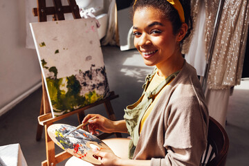 Young female creative pretty african american artist paints picture with oil paints in art studio