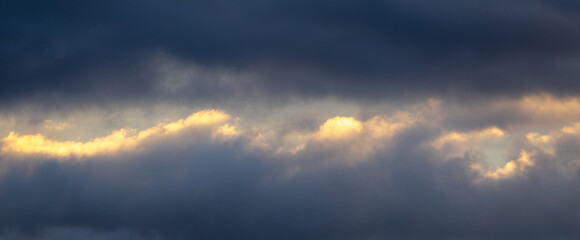Naklejka na ściany i meble A patch of sky illuminated by the evening sun shines through the dark blue clouds