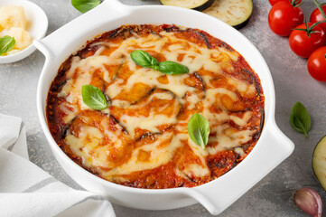 Eggplant casserole with cheese and tomato sauce in a white baking dish on a gray concrete background with ingredients for cooking. Vegetarian healthy food.