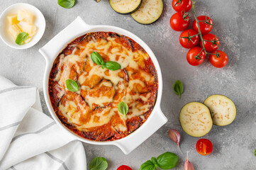 Eggplant casserole with cheese and tomato sauce in a white baking dish on a gray concrete background with ingredients for cooking. Vegetarian healthy food.