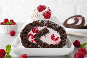 Chocolate roll cake with cream and fresh raspberries on a white plate on a gray background. Delicious holiday dessert.