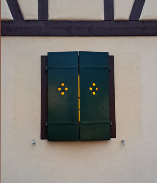 Dunkelgr&uuml;ner Fensterladen sticht farblich von der wei&szlig;en Wand ab. Durch die L&ouml;cher und Ritzen scheint warmes Licht. &Uuml;ber dem Fenster sind schwarze Balken eines frisch renovierten Fachwerks zu sehen.  
