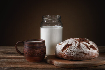 fresh bread on a cutting board with a glass jar and a clay mug with milk on a brown background with a gradient. simple rustic still life. side view