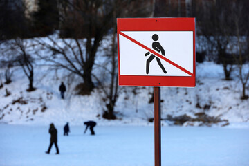 Sign 'Access to the ice is prohibited' in the snowy park. People walking on frozen lake surface in...