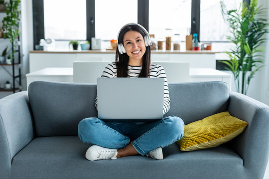 Beautiful Young Woman Working With Her Laptop While Listening To Music With Earphones Sitting On A Couch At Home.