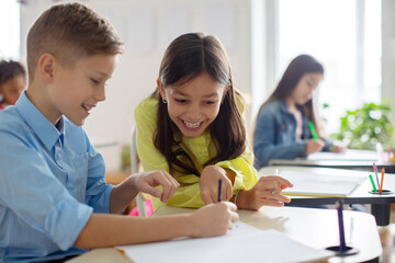 Schoolgirl sitting at table near boy classmate and pointing in copybook of friend with finger, enjoying learning process in classroom