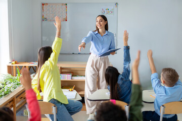 Friendly female teacher in classroom teaches elementary school students, kids raising hands to teacher aasks questions to small students sitting at their desks