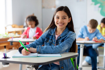 Smiling junior school girl sitting at desk in classroom, writing in notebook, posing and looking at camera. Group of diverse classmates studying on background
