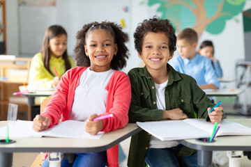 Happy black schoolboy and schoolgirl looking at camera and smiling, sitting at desks with other children on background