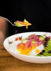 Egg yolk dripping from a fork while someone is eating a fried egg and avocado salad
