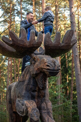 Fototapeta na wymiar A father and son hold hands high on the antlers of a large wooden moose sculpture. Pine treetops in the background