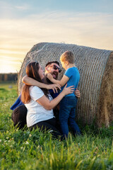 Cheerful young family hugging tightly and joking near haystack in summer green fields