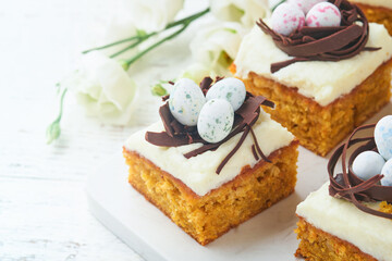 Easter carrot cake bars decorated with chocolate nest and chocolate candy eggs blossoming cherry or apple flowers on rustic light wooden backgrounds. Easter holiday meal. Traditional Easter food.