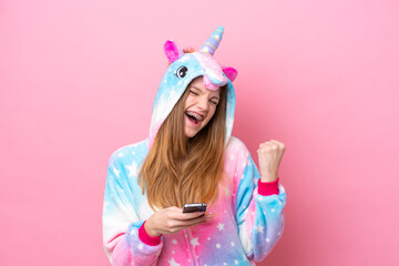 Teenager Russian girl with unicorn pajamas isolated on pink background with phone in victory...