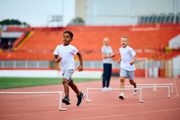 Obraz premium Black kid running on track with hurdles during sports training at stadium.