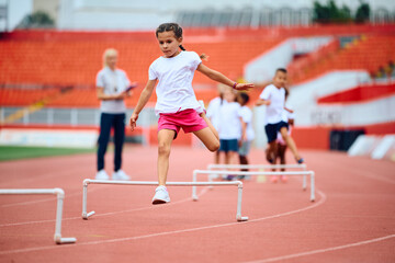 Obraz premium Little girl jumping over obstacles while running during exercise class at stadium.