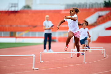 Obraz premium Black girl running on hurdling track at stadium.